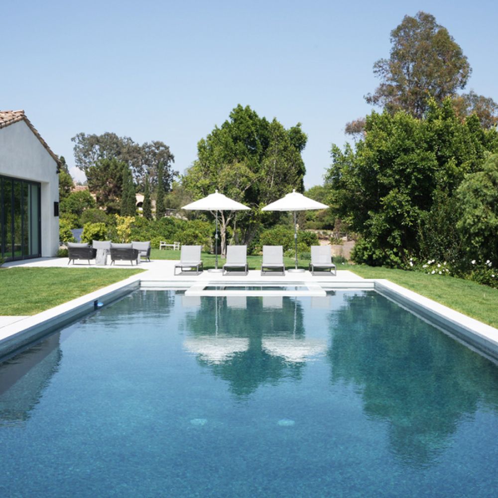 <span class="uk-h4">Modern backyard pool with lounge chairs, umbrellas, and Spanish-style home surrounded by lush landscaping on a sunny day.</span>