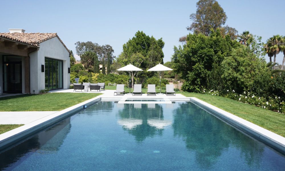 <span class="uk-h4">Modern backyard pool with lounge chairs, umbrellas, and Spanish-style home surrounded by lush landscaping on a sunny day.</span>