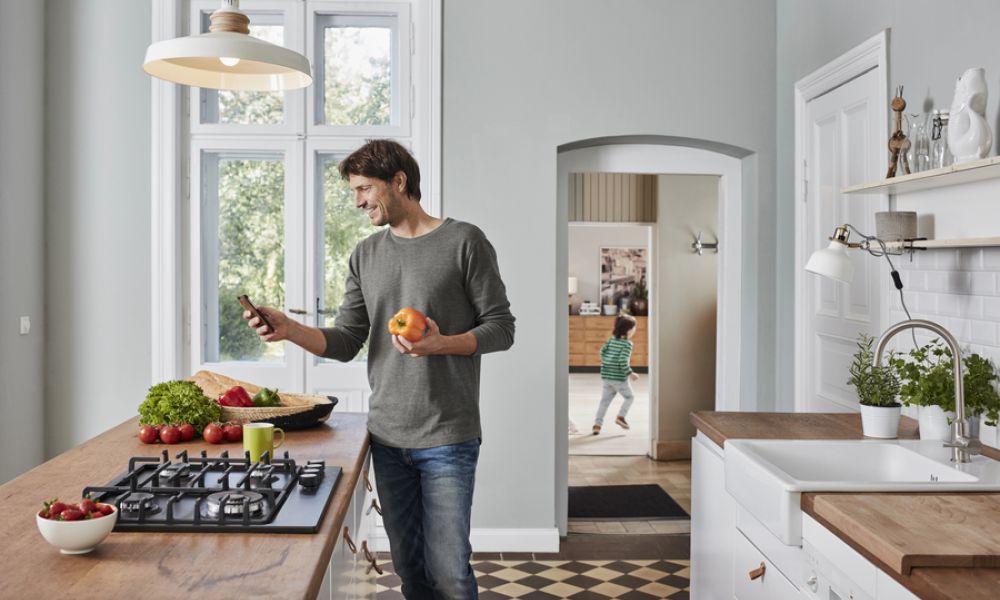 <span class="uk-h4">Man checking his phone while prepping food at a sunny kitchen island, with his kid playing in the background.</span>