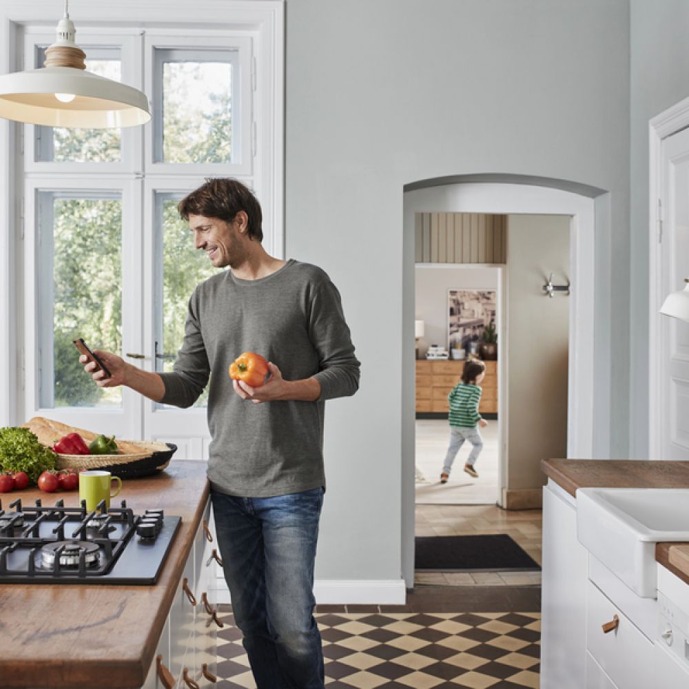 <span class="uk-h4">Man checking his phone while prepping food at a sunny kitchen island, with his kid playing in the background.</span>