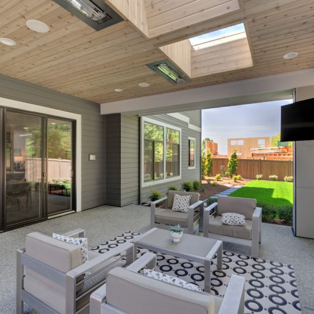 <span class="uk-h4">Covered outdoor patio with modern gray seating, skylights, ceiling heaters, a mounted TV, and a view of a landscaped backyard with grass and shrubs.</span>
