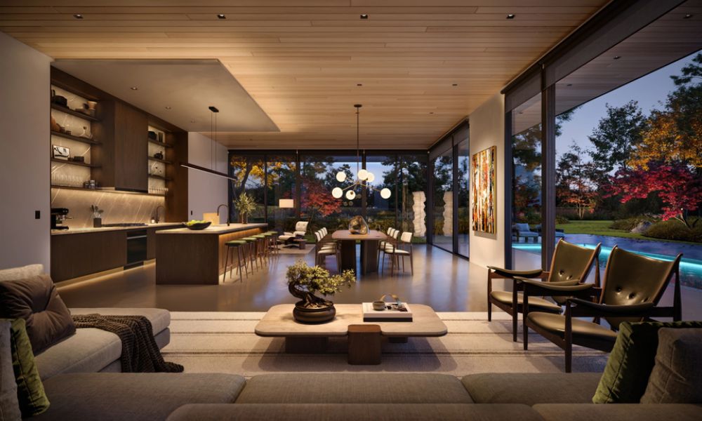 <span class="uk-h4">Modern open-plan great room at dusk with warm lighting, walnut kitchen, and glass walls opening to a pool and garden featuring fall foliage.</span>